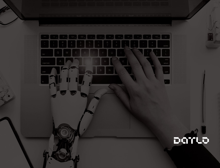 Imagem de duas mãos digitando em um notebook, uma delas sendo de um robô e a outra de uma pessoa