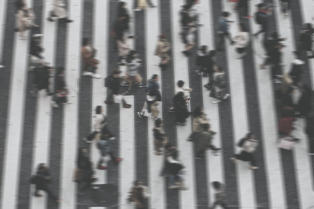 Pessoas andando em uma faixa de pedestres, capturadas em movimento, com um efeito de desfoque, dando a sensação de dinamismo e fluxo urbano.