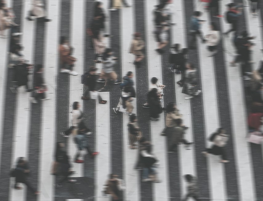Imagem colorida, tirada de cima com desfoque, a imagem é composta por uma faixa de pedestre com pessoas atravessando.