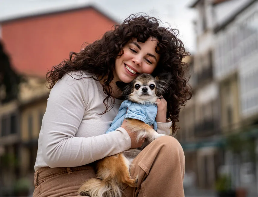 Imagem colorida composta por uma mulher abraçando seu cachorro, com o fundo de uma cidade desfocada, a imagem ilustra pontos e atrações turísticas que permitem a entrada de animais