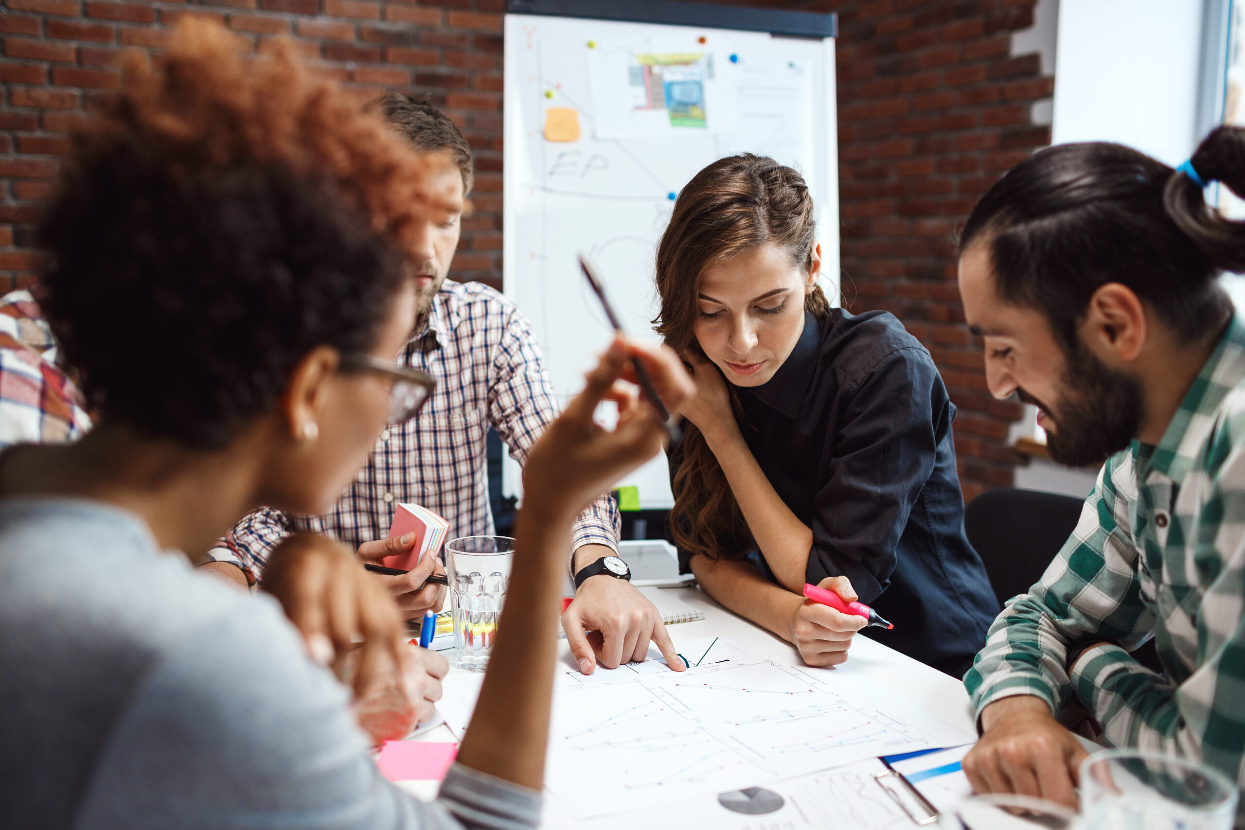 Imagem ilustrativa para representar pessoas executando a estratégia de go-to-market em uma empresa. Na imagem, já quatro pessoas, dois homens e duas mulheres! Ao fundo existe um quadro na vertical com papéis coloridos e brancos pregados, parede de tijolos. Na mesa há marca textos, blocos de papel, folhas, canetas, copo de água. As quatro pessoas estão sorrindo e analisando um papel.